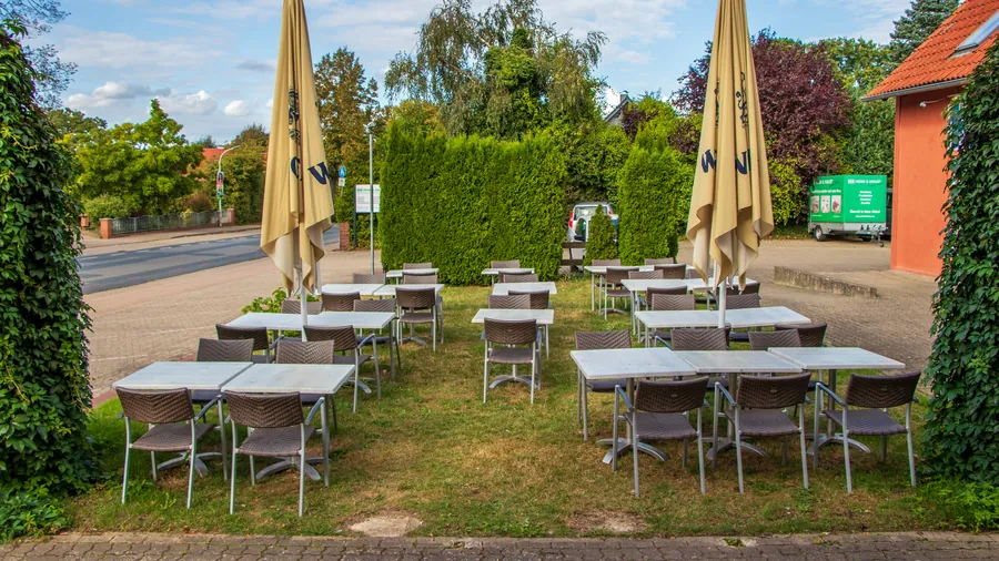 Tische und Stühle auf der Wiese neben dem Gasthaus, zwei Sonnenschirme, dahinter Hecken